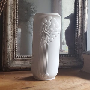 Może przedstawiać: Biały ceramiczny wazon z wypukłym wzorem kwiatowym. Wazon jest wysoki i smukły, z zaokrągloną podstawą i lekko rozszerzającym się wierzchołkiem.