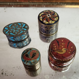 Op de afbeelding: Vier kleine, ronde, decoratieve doosjes met ingewikkelde beschilderde ontwerpen. De doosjes zijn gemaakt van metaal en hebben een glanzende afwerking. De ontwerpen omvatten een blauw en turquoise bloemenpatroon, een rood en geel bloemenpatroon, een rood en geel vogelpatroon en een blauw en rood pauwenpatroon.
