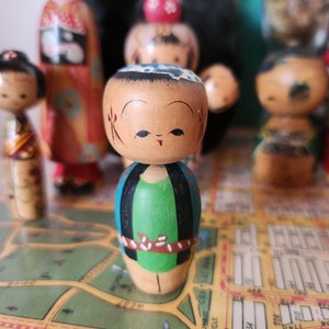 May include: A collection of wooden dolls with painted faces and colourful clothing. The dolls are arranged on a map with a green background. The dolls are traditional Japanese kokeshi dolls.