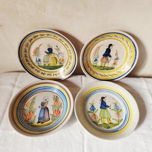 Könnte beinhalten: Vier antike Keramikplatten mit handgemalten Volkskunstmotiven. Jede Platte zeigt eine zentrale Figur in traditioneller Kleidung, umgeben von floralen Motiven und einem dekorativen Rand in Blau, Gelb und Orange. Die Teller sind auf einer weißen Oberfläche angeordnet.
