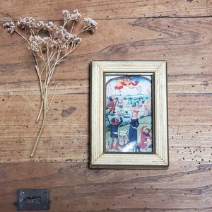 Op de afbeelding: Een ingelijst vintage-illustratie die een middeleeuwse scène met mensen in kleurrijke kleding afbeeldt. De illustratie is tegen een achtergrond van een stad met gebouwen en een blauwe lucht. De lijst is wit met een gouden rand.