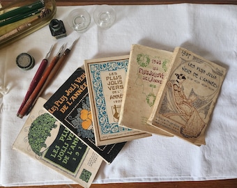 May include: A collection of vintage books with the title "Les Plus Jolis Vers de l'Année" in French. The books are bound in beige paper with decorative floral designs and feature the year of publication on the cover. The books are arranged on a white linen tablecloth.
