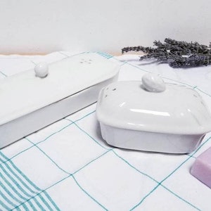 Peut inclure: Deux savonnettes en céramique blanche avec couvercles. La plus grande est rectangulaire et la plus petite est carrée. Les deux savonnettes ont un petit bouton sur le couvercle. Les savonnettes sont posées sur une serviette à carreaux blancs et verts.