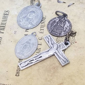 May include: A set of four silver-toned religious charms, including a crucifix, a round charm with a woman in a robe, and two oval charms with women in robes. The charms are on a brown paper background with text in French.