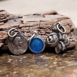 May include: Three silver charms with religious themes. The charms are on a wooden surface. One charm features a woman's face, another has a blue enamel design, and the third is a rose.