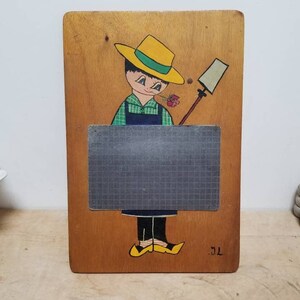 Könnte beinhalten: Ein vintage Holztafel mit einer handgemalten Illustration einer Person mit einem gelben Hut, die eine Blume hält. Die Tafel hat eine graue Oberfläche mit einem Gittermuster und die Initialen "J.L." sind in der rechten unteren Ecke gemalt.