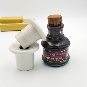 May include: A vintage glass bottle of violet ink with a cork stopper. The bottle has a label that reads "Encre Violette Marque Dagron Paris". The bottle is sitting on a white ceramic inkwell.