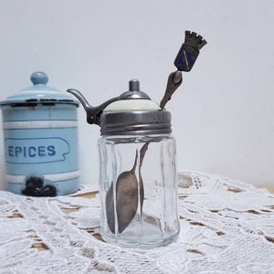 Puede incluir: Un dispensador de azúcar de vidrio transparente con una tapa plateada y una cuchara plateada con una parte superior decorativa. El dispensador está sentado sobre una servilleta de encaje blanco.