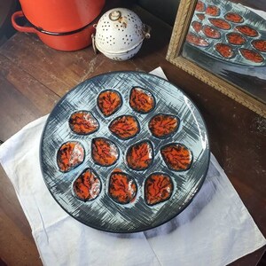 May include: A round ceramic serving platter with a blue and gray background and 12 orange and black leaf-shaped indentations.