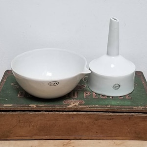 May include: A white porcelain bowl and funnel, both with a black stamp, sitting on top of a wooden box with green paint and the words "De l'eau au peuple".