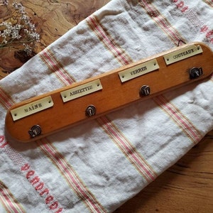 Può includere: Un portautensili in legno con cinque ganci ed etichette metalliche per organizzare gli utensili da cucina. Le etichette recitano "Mains", "Assiettes", "Verres", "Couteaux".