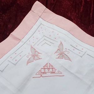 Può includere: Un copri cuscino in lino bianco con un bordo rosa e farfalle ricamate e un monogramma. Il ricamo è realizzato in un delicato motivo floreale.