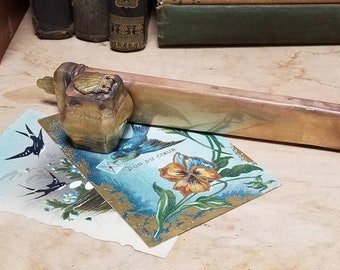 May include: A long, rectangular, brass doorstop with a decorative, ornate, gold-colored top. The doorstop is resting on a vintage postcard with a floral design and the text "Don du Coeur".