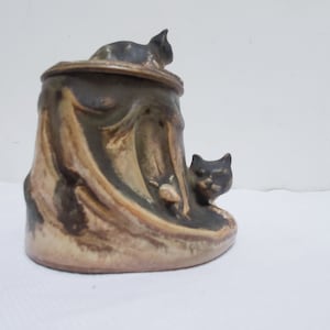 May include: A ceramic jar with a tree trunk design and two black cat figurines. The jar has a brown, tan, and white glaze. The cats are perched on the top of the jar and on the side.