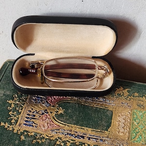 Può includere: Una custodia in pelle nera con occhiali da lettura pieghevoli dorati all'interno. La custodia è aperta e gli occhiali sono visibili. La custodia è appoggiata su una superficie con un motivo verde e dorato.