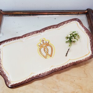 May include: A white ceramic serving tray with a brown rim and a gold design of two intertwined hearts with a crown on top. A small bouquet of white flowers is on the tray.