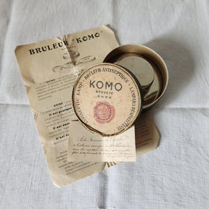 May include: An antique Komo Brevete antiseptic lamp with a round tin container and a paper label. The label features the text "BRULEUR KOMO" and "ANTISEPTIC LAMP". The image also includes a vintage paper with text.