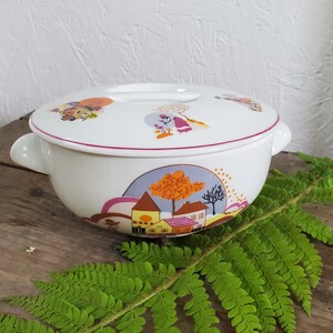 Op de afbeelding: Witte keramische ovenschaal met deksel. De schaal heeft een roze rand en een kleurrijk ontwerp met huizen, bomen en mensen. Het deksel heeft een handvat en een vergelijkbaar ontwerp.
