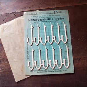 Peut inclure: Une feuille de lettres de monogramme en feutre blanc pour la broderie. Les lettres sont en écriture cursive et sont disposées en rangées sur une grille bleue. La feuille est étiquetée "MonoGramme a Broder" et "La Douzaine 6 Fr. 00".