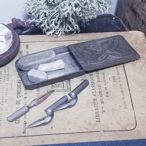 May include: A vintage metal tin with a butterfly design on the lid. The tin contains two small rectangular pieces of white chalk. The tin is sitting on a book with French text. There are also two silver metal nib pens on the book.