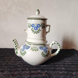 Può includere: Una caffettiera in ceramica bianca con coperchio abbinato e sezione superiore separata. La caffettiera è decorata con motivi floreali blu e dettagli verdi. Anche il manico e il beccuccio sono rifiniti in verde.