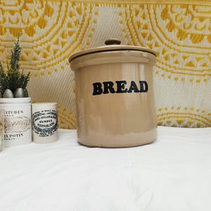 May include: A tan ceramic bread bin with a lid and the word "BREAD" in black lettering. Several small decorative items are arranged to the left, including candles, a small vase with greenery, and a small jar.