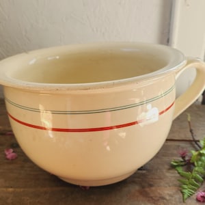 May include: A white ceramic bowl with a green and red striped design. The bowl has a wide, rounded base and a handle on one side.