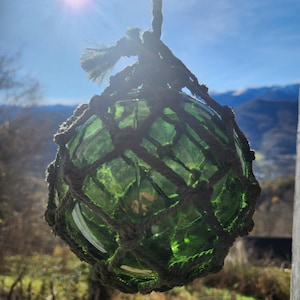 Op de afbeelding: Een groene glazen boei, opgehangen aan een geweven touwnet en een dik touw. De glazen bol is zichtbaar tegen een helderblauwe lucht en een zonnevlam. De boei is een decoratief nautisch item.