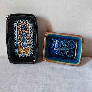 May include: Two ceramic rectangular dishes with decorative designs. The larger dish is black with a floral design in shades of blue, red, and yellow. The smaller dish is brown with a blue and green design featuring a bird and swirls.