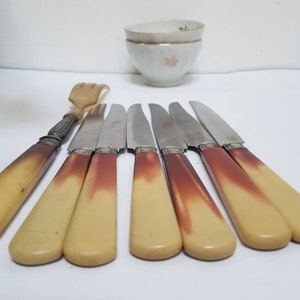 Peut inclure: Ensemble de huit couteaux vintage avec des manches de couleur ivoire et des accents argentés. Les couteaux ont des lames en acier inoxydable et sont disposés en rangée sur une surface blanche.