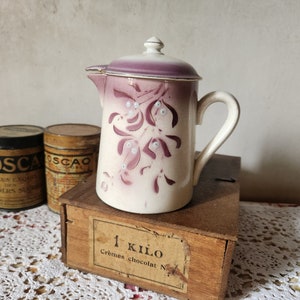 Peut inclure: Un pichet en céramique blanche avec un couvercle violet et un motif floral. Le pichet a une poignée et un bec verseur. Il est posé sur une boîte en bois avec une étiquette qui dit "1 Kilo Cremes chocolat N".