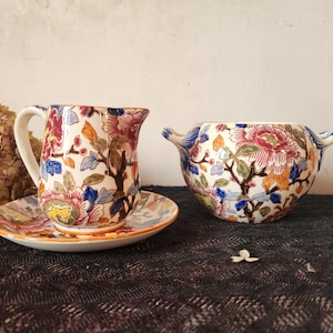 Puede incluir: Un juego vintage de cerámica que incluye jarra, platillo y cuenco. Las piezas presentan un estampado floral con flores rojas, azules y naranjas sobre un fondo crema. La jarra tiene un asa y el cuenco tiene dos asas pequeñas. El platillo es redondo.