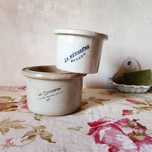 May include: Two antique, off-white ceramic jars with blue text, one stacked on top of the other. The top jar reads "LA MENAGERE" and the bottom jar reads "Les Conseros". A soap dish with a bar of soap is in the background.
