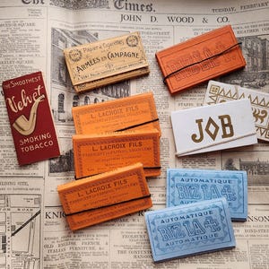 Pode incluir: Uma variedade de embalagens de tabaco e cigarros vintage, incluindo caixas de "Velvet Smoking Tobacco", "L. Lacroix Fils" e "Riz La". Os itens estão dispostos em um fundo de jornal, com texto e gráficos.