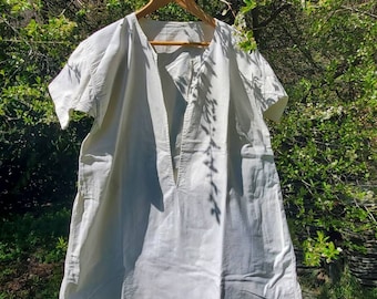 May include: A white cotton tunic with a V-neck and short sleeves hanging on a wooden hanger. The tunic is hanging in front of a green bush with white flowers.