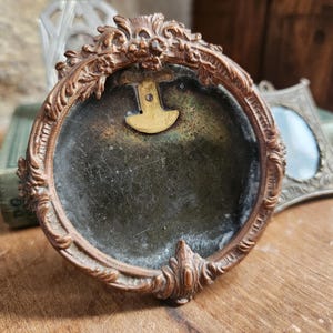 May include: A small, ornate, oval-shaped, brass picture frame with a dark, aged, glass insert. The frame has a decorative, floral design and a brass hinge on the back.