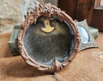 May include: A small, ornate, oval-shaped, brass picture frame with a dark, aged, glass insert. The frame has a decorative, floral design and a brass hinge on the back.