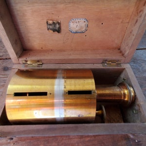 May include: A brass cylinder with two slots in a wooden box. The box has a label with the text "'63" on it.