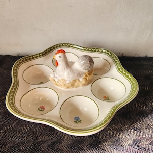Pode incluir: Um porta-ovos de cerâmica com uma figura de galinha branca no centro. O porta-ovos tem seis reentrâncias circulares e é decorado com uma borda verde e pequenos detalhes florais. A galinha tem uma crista vermelha e penas cinzentas.