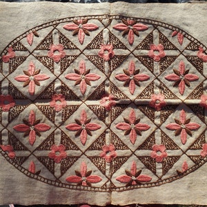May include: A beige fabric oval with a brown and pink embroidered design. The design features a repeating pattern of flowers and leaves.