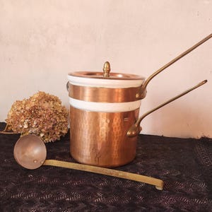 May include: A copper and white cooking pot with a lid and two handles, and a matching ladle. The pot has a hammered texture and a brass handle. The ladle has a round bowl and a long handle. A dried flower is in the background.
