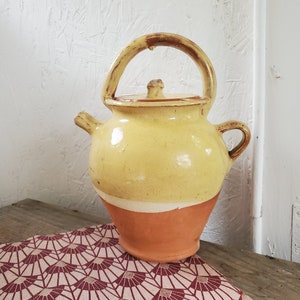 May include: A yellow ceramic pitcher with a brown handle and a white rim. The pitcher is sitting on a red and white patterned surface.