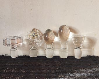 May include: Five clear glass bottle stoppers of varying shapes and designs. The stoppers include a rectangular, a floral, an oval, a teardrop, and a ribbed design. The stoppers are displayed on a black lace surface.