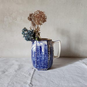 May include: A ceramic pitcher featuring a blue and white transferware pattern depicting a cathedral with tall spires. The pitcher has an angular handle and contains a small arrangement of dried brown and blue flowers extending from the top opening.