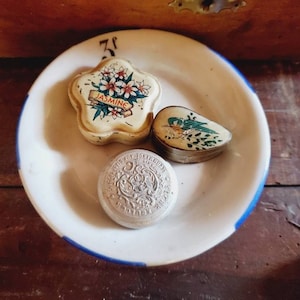 Può includere: Tre scatole vintage in metallo per bigiotteria su un piatto bianco con un bordo blu. Una scatola ha la forma di un fiore e ha la parola "Jasmine" stampata su di essa. Un'altra scatola ha la forma di un cuore e ha un disegno di un uccello. La terza scatola è rotonda e ha un disegno floreale.