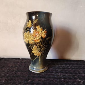 Op de afbeelding: Een hoge, donkere vaas met een uitlopende bovenkant en een brede basis. De vaas is versierd met gele en oranje bloemmotieven. De achtergrond is neutraal van kleur en de vaas staat op een donkere ondergrond.