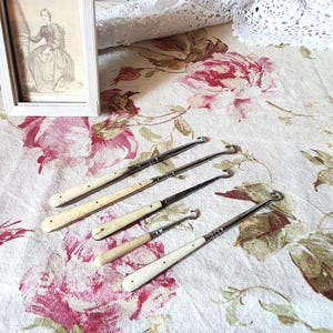 May include: Five antique crochet hooks with ivory-colored handles and metal hooks are arranged on a floral patterned fabric. A framed portrait of a woman in a dress is in the background. The fabric has a pink and cream floral pattern.