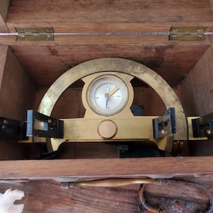 May include: A brass compass and protractor in a wooden box. The compass has a white face with black markings and a blue needle. The protractor is marked in degrees.