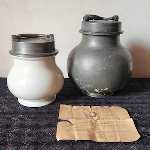 May include: Two antique jars with metal lids. One is white ceramic, the other is a dark gray metal. A torn, aged paper with text lies in front of them. The jars are likely for storage.