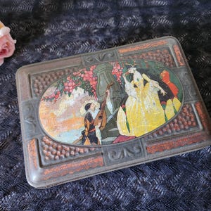 May include: Vintage rectangular metal tin with an oval-shaped illustration on the lid. The illustration depicts figures in yellow and white gowns, a man playing a lute, and a floral background. The tin has a textured, aged appearance.